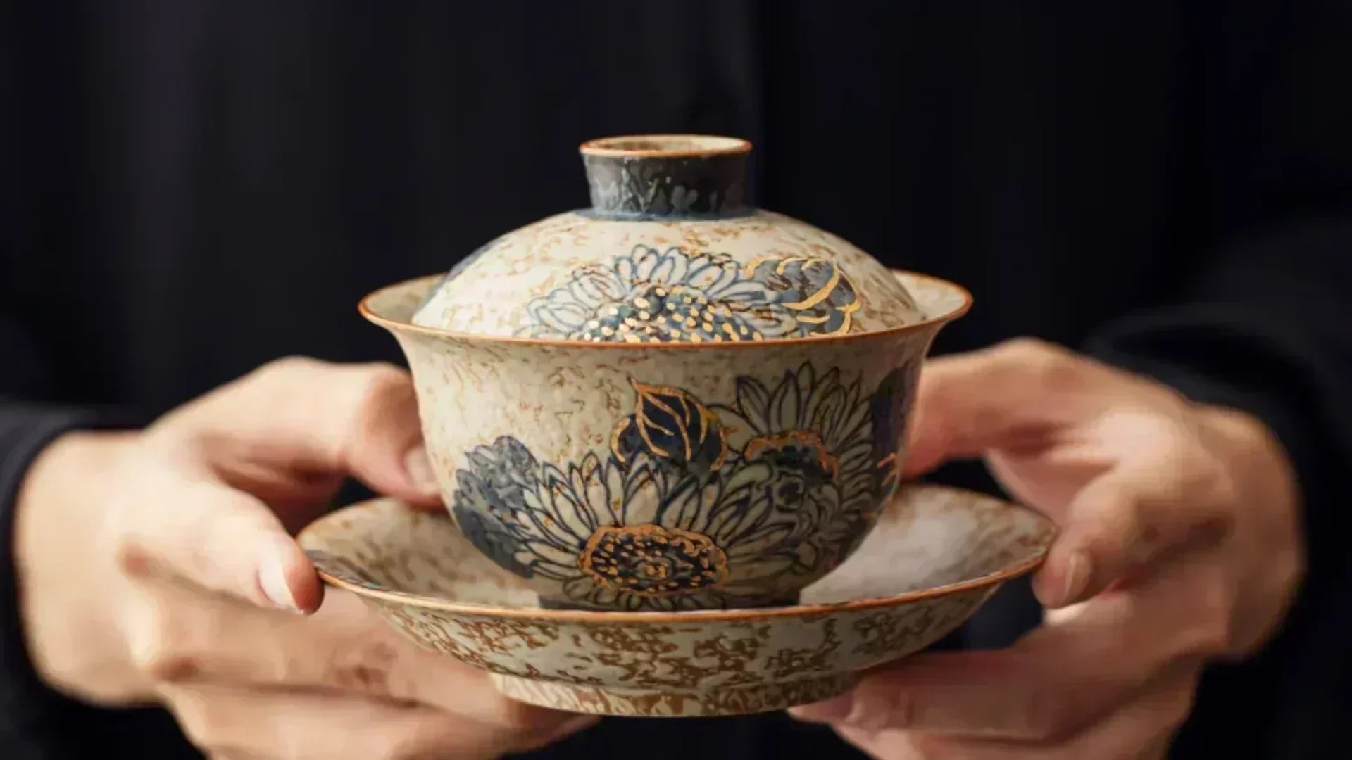 Two hands holding a decorative porcelain gaiwan, a traditional Chinese tea brewing vessel, during a tea ritual.