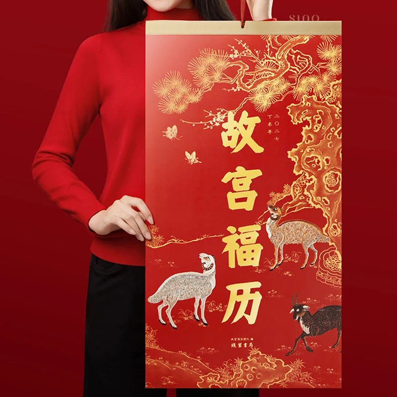 2027 Year of the Fire Goat wall calendar held against a red background, featuring golden Chinese characters and auspicious goat illustrations for Chinese New Year decor.