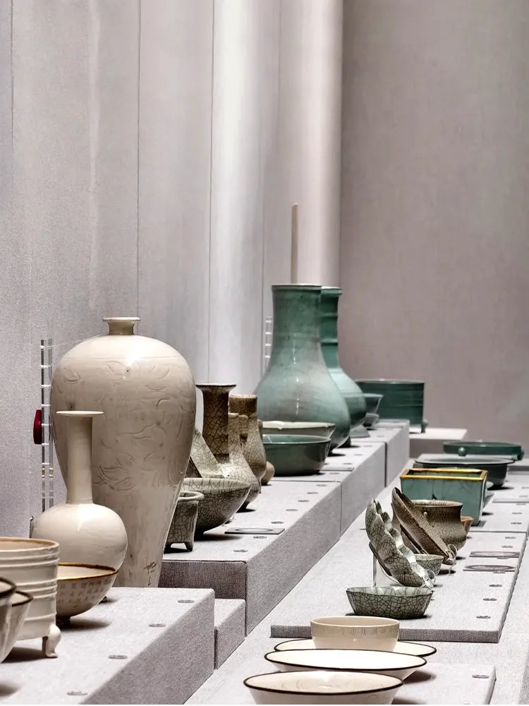Display of Chinese porcelain: white carved vases, celadon bowls, and crackleware in a museum setting.