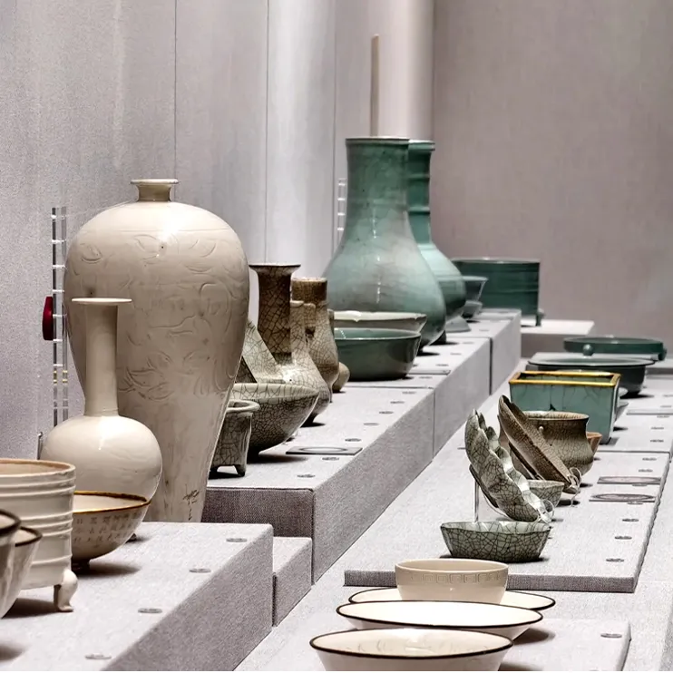 Display of Chinese porcelain: white carved vases, celadon bowls, and crackleware in a museum setting.