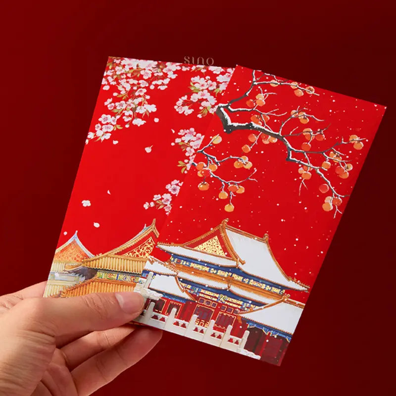 Hand holding two traditional Chinese red packets decorated with pink blossoms, snow-covered persimmon branches, and colorful Chinese palace rooftops.