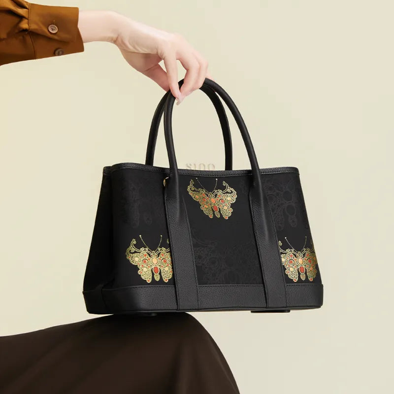 Seated model displaying the luxury black Song brocade gold butterfly motif tote bag on the knees, holding the handles.