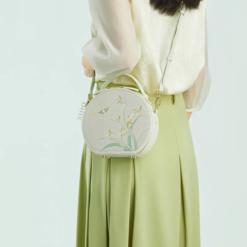 Side view of model wearing the Chinoiserie Luxury Song Brocade Phalaenopsis Orchid Round Bag crossbody, holding the strap with one hand.