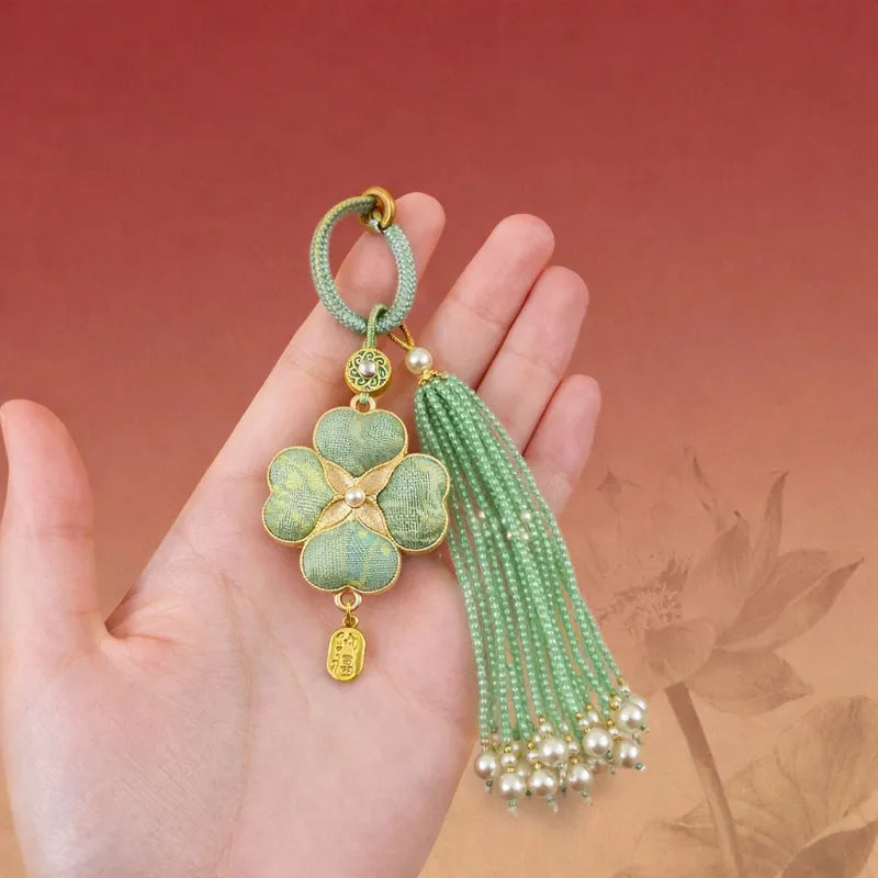 Green Chinoiserie Song Brocade auspicious four-leaf clover tassel bag charm displayed on a hand, with pearl tassels and gold-tone trim.