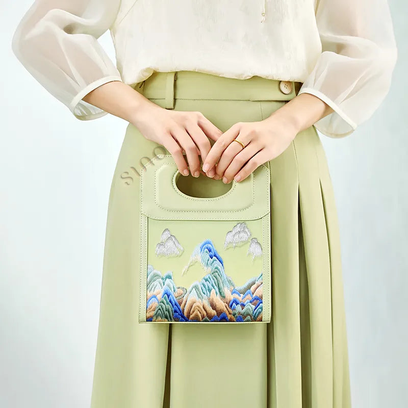 Front view of the Chinoiserie Mountains Painting hand-embroidered leather handle bag with blue and green landscape embroidery.