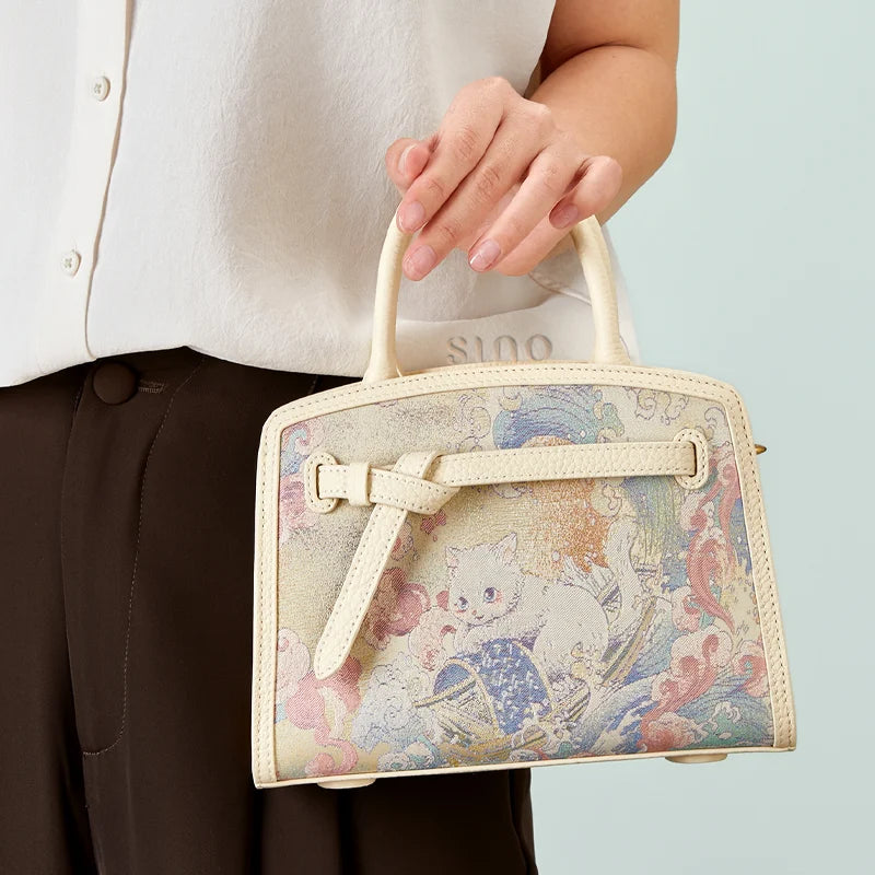 Model carrying the Luxury Song Brocade Brave Voyage Leather Top Handle Bag by its double handles, highlighting the artistic cat and voyage design.