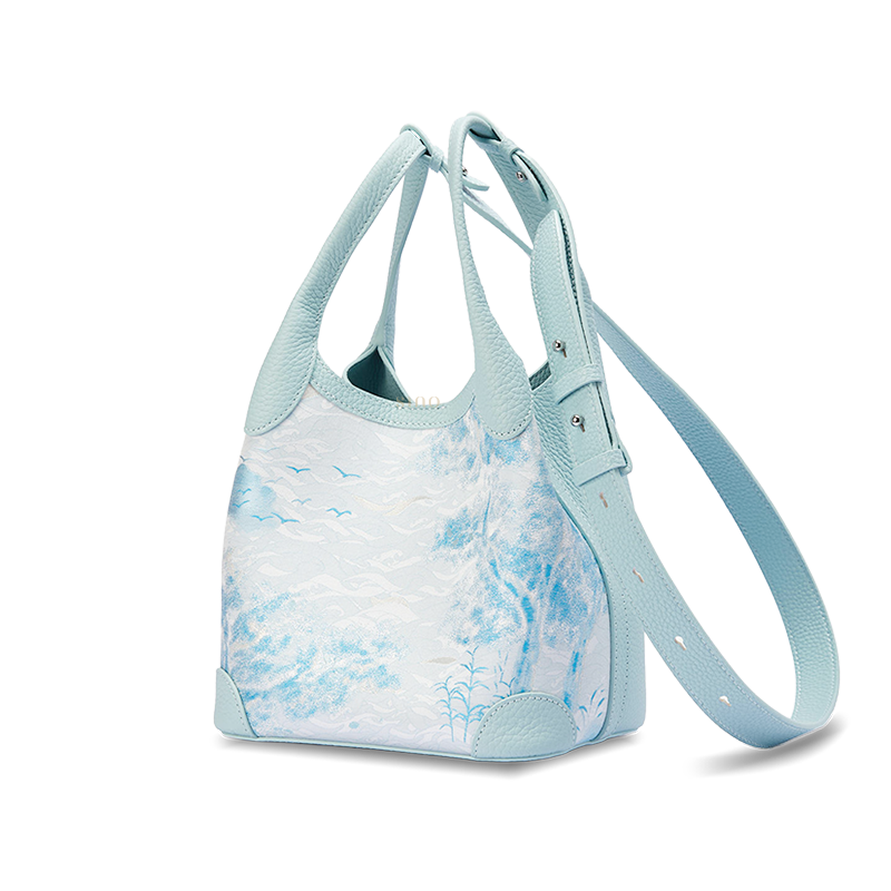 Angled view of the Luxury Song Brocade Misty Mountain Forest Leather Basket Tote Bag with adjustable long strap, showing the pale blue leather side panel and woven landscape pattern.