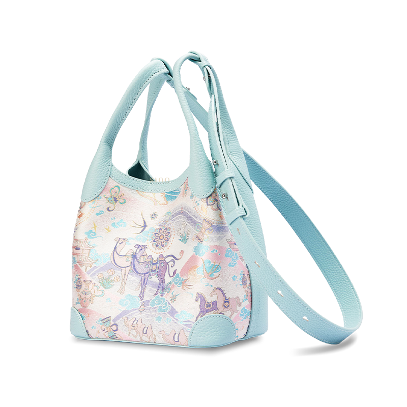 Angled view of the Song Brocade Silk Road Leather Basket Tote Bag with adjustable long strap, showing the light blue leather side panel and woven Silk Road pattern.