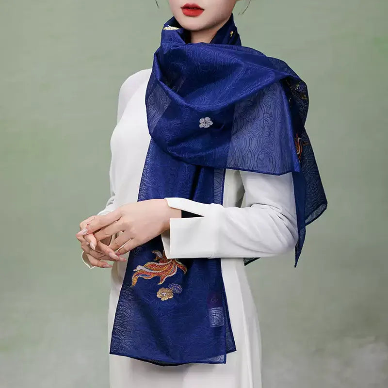 Model wearing the Nanjing Yunjin silk shawl loosely wrapped around the neck over a white dress, showing its translucent border and colorful woven fish.