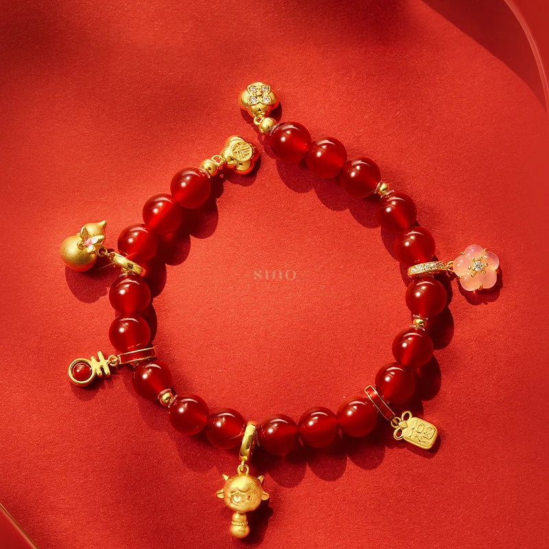 Flat lay of the 2027 Year of the Goat red agate bracelet featuring polished red beads, gold-tone lucky charms and a pink blossom pendant against a festive red background.