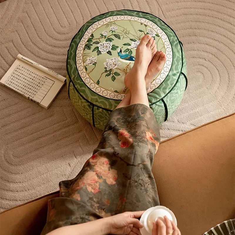 Full top-down view of a model on a sofa with their feet resting on the chinoiserie jade green floor pouf used as a footrest.