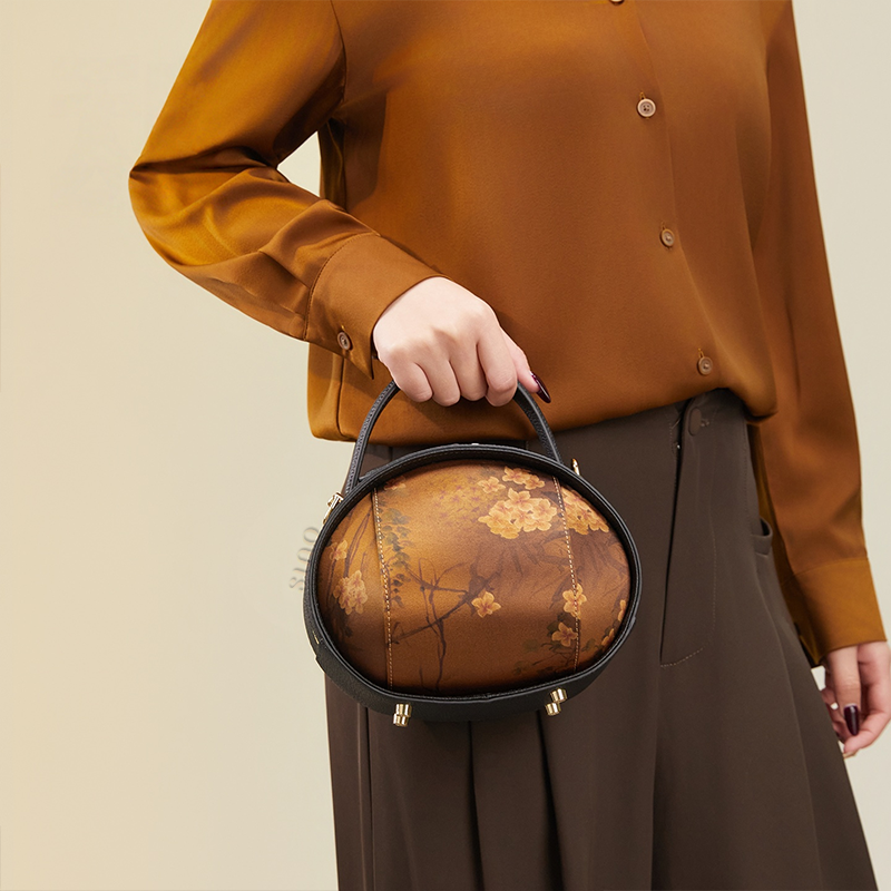 Model standing left holding black Xiangyunsha handbag (Plum Blossom Print) with one hand.