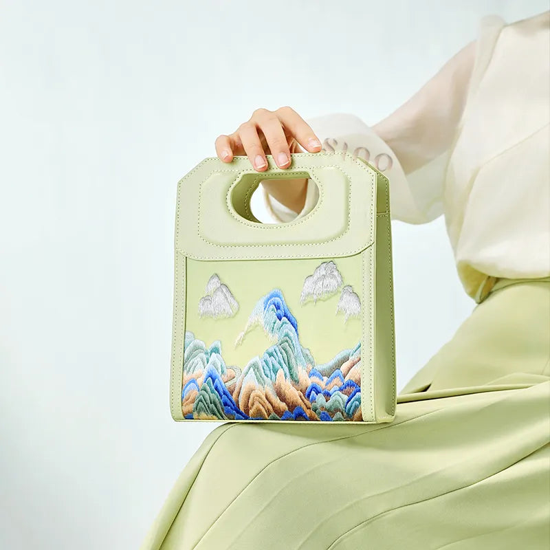 Model holding a Chinoiserie Mountains Painting hand-embroidered leather handle bag in light green, featuring blue mountain embroidery.