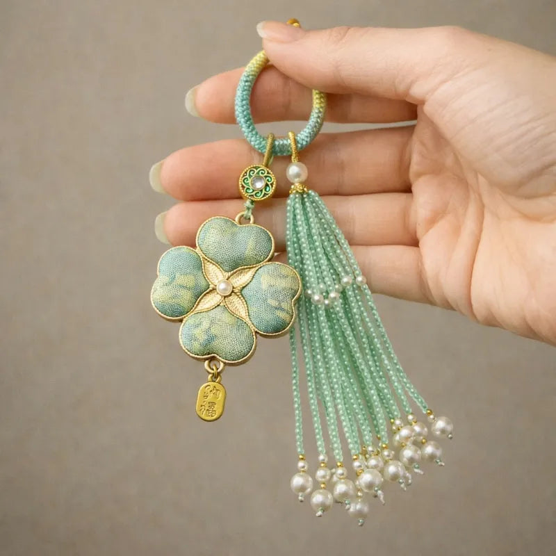 Hand holding a green Chinoiserie Song Brocade auspicious four-leaf clover tassel bag charm with pearl tassels and gold-tone details.