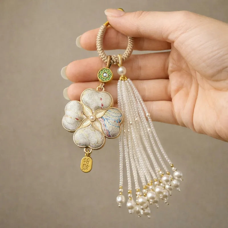 Hand holding a pink Chinoiserie Song Brocade auspicious four-leaf clover tassel bag charm with pearl tassels and gold-tone details.