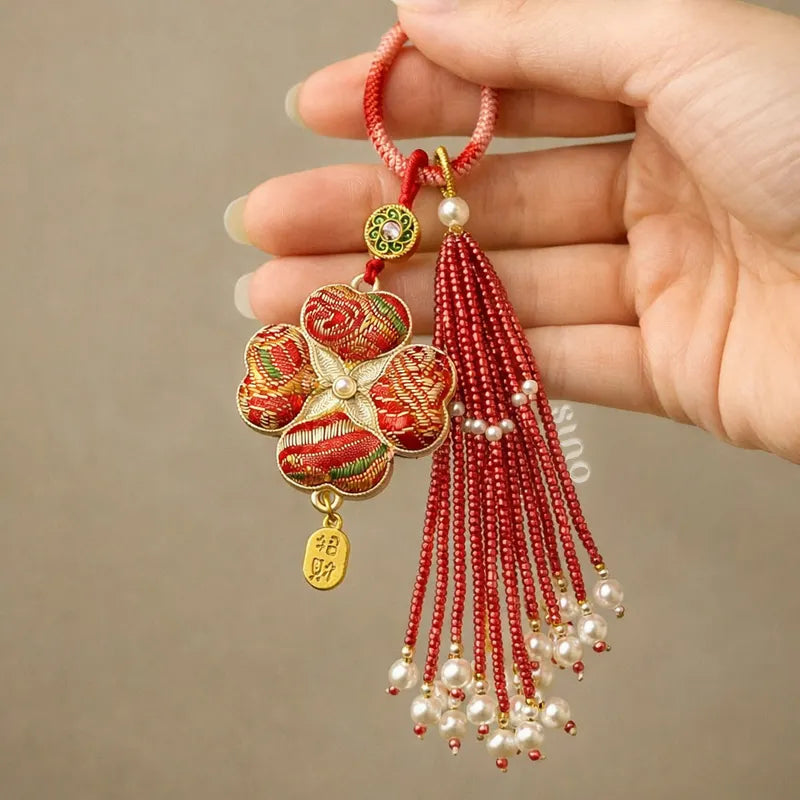 Hand holding a red Chinoiserie Song Brocade auspicious four-leaf clover tassel bag charm with pearl tassels and gold-tone details.