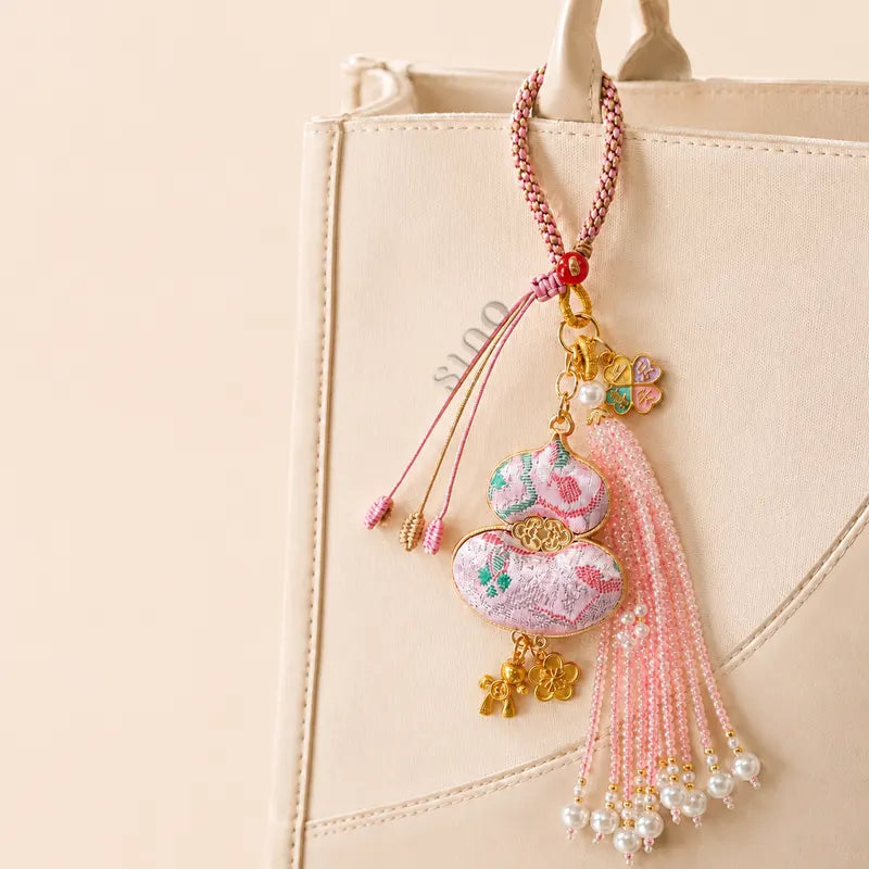 Pink Chinoiserie Song Brocade auspicious hulu pearl tassel bag charm hanging on a beige handbag, featuring pearl tassels and gold-tone charms.
