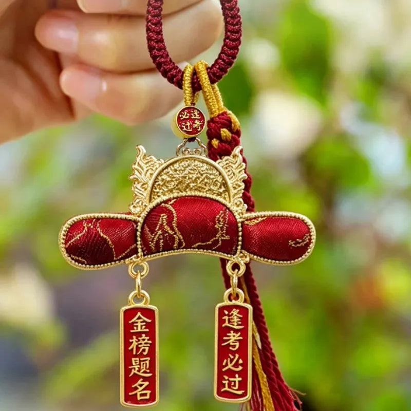 Handheld red Nanjing Yunjin brocade Imperial Scholar Hat bag charm with gold-toned trim, braided cord, and auspicious exam blessing pendants.