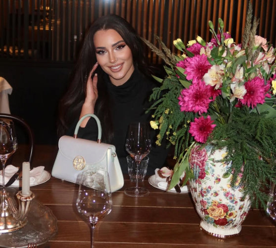@lizastasia in a black turtleneck crop top and leather pants, holding a white floral handbag, standing in a restaurant.