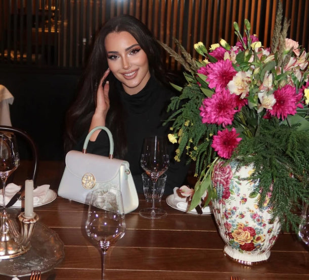 @lizastasia in a black turtleneck crop top and leather pants, holding a white floral handbag, standing in a restaurant.