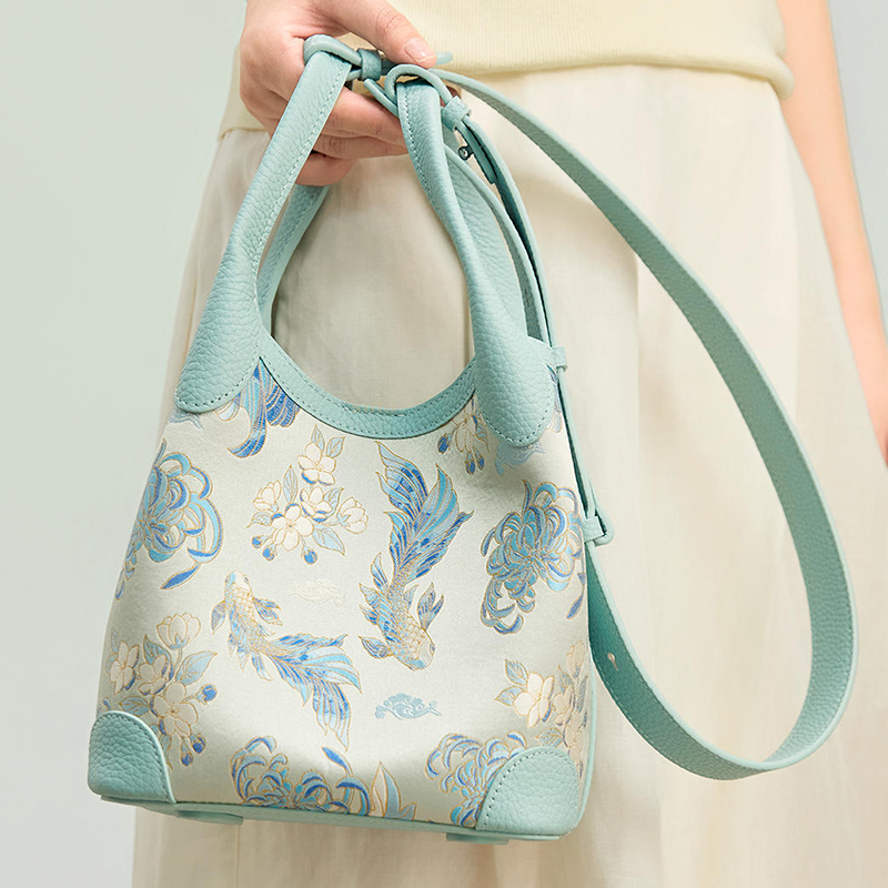 Handheld model view of the Luxury Shu Brocade Golden Fish & Flower Leather Basket Tote Bag, highlighting the golden fish motif, floral brocade pattern, and shoulder strap.