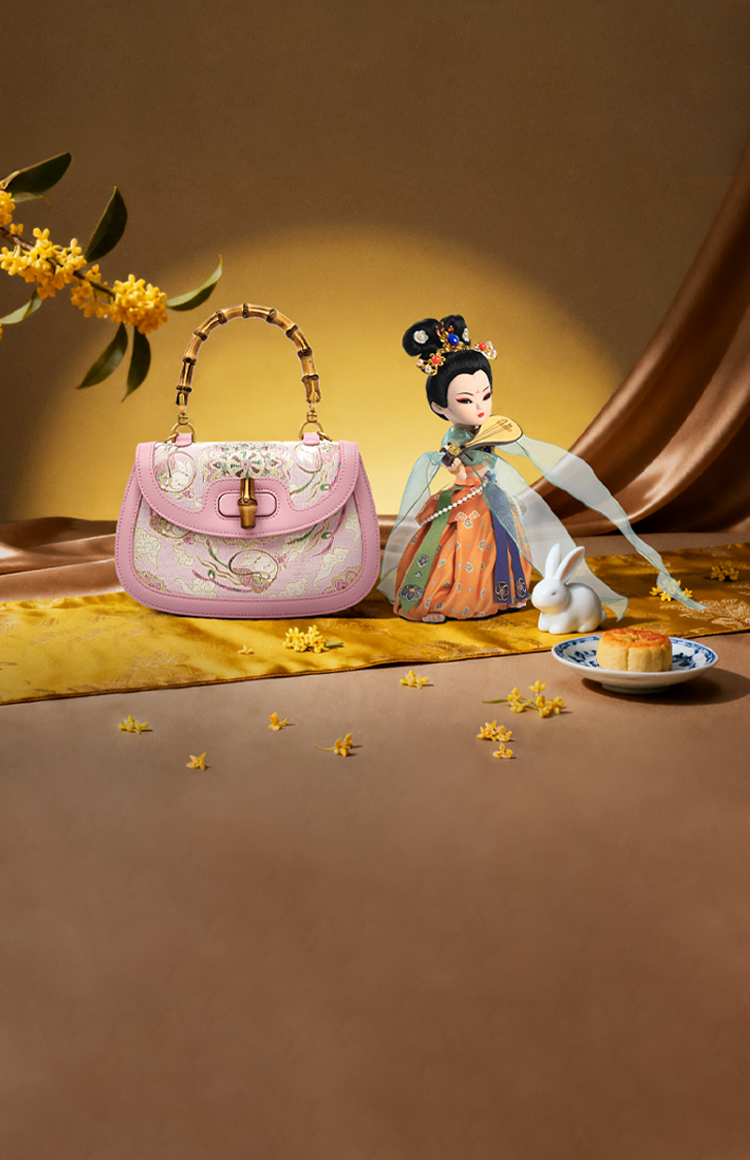 A traditional Dunhuang Flying Apsara doll in embroidered attire beside a pink bamboo-handle bag, with osmanthus flowers, a white rabbit, and a mooncake on a golden silk backdrop for Mid-Autumn Festival.