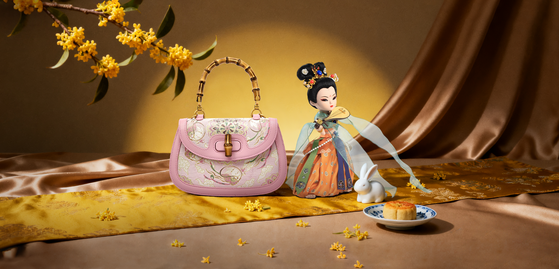A traditional Dunhuang Flying Apsara doll in embroidered attire beside a pink bamboo-handle bag, with osmanthus flowers, a white rabbit, and a mooncake on a golden silk backdrop for Mid-Autumn Festival.