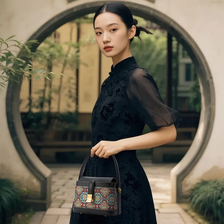 A woman in a black embroidered dress holds a compact handbag with a vibrant Song Brocade pattern, standing in a classical Chinese garden archway.