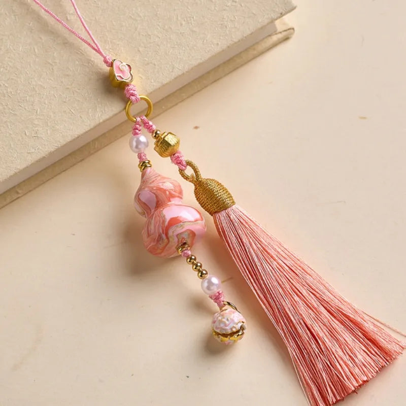 Pink Daqi lacquer Hulu tassel bag charm with a marbled finish, pearl accents, gold-toned details, and a long pink tassel displayed on a beige background.