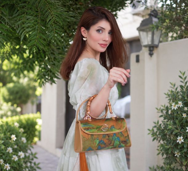 SinoCultural x @hinaashh featuring a Xiangyunsha silk bamboo-handle handbag with A Thousand Li of Rivers and Mountains artwork, styled with a pale green dress outdoors.