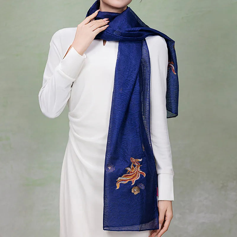 Model styling the blue Nanjing Yunjin brocade shawl as a long silk scarf over a white dress, showing the colorful fish motif near the hem.