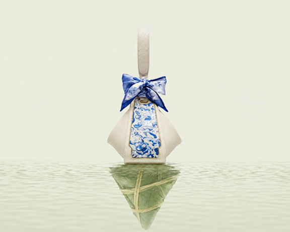 A cream SANLAN embroidered Zongzi bag with a blue silk bow reflected on calm water.