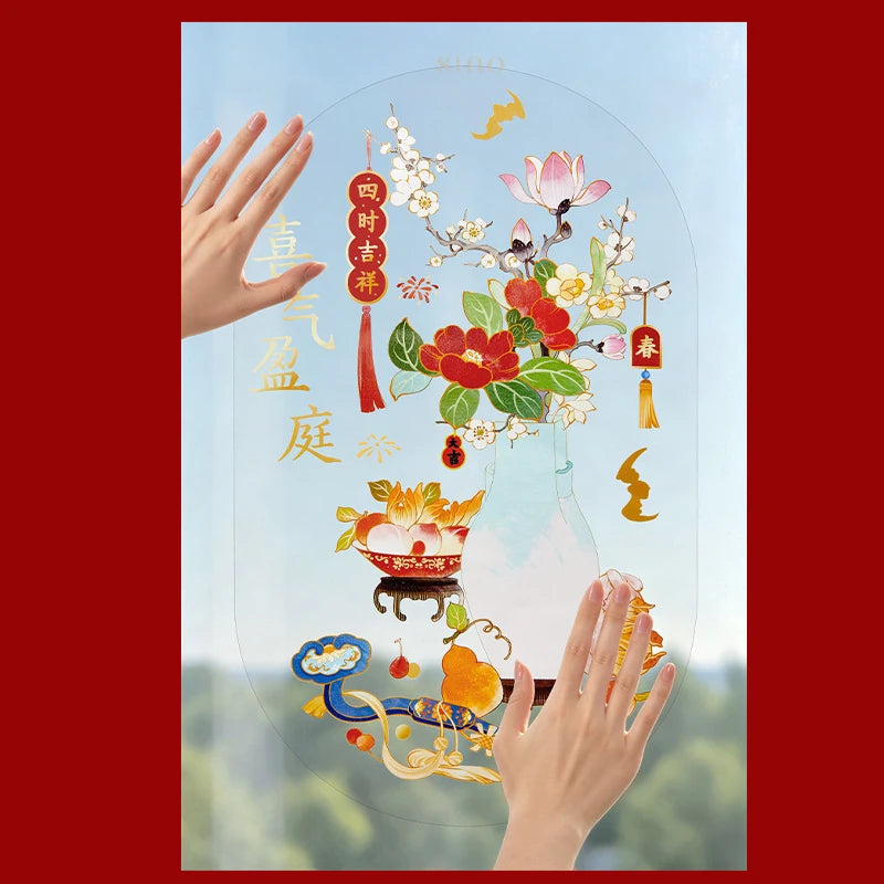 Hands positioning a Year of the Goat window cling featuring a pale blue vase, red camellias, plum blossoms, auspicious fruits, and gold Chinese lettering against a bright window.